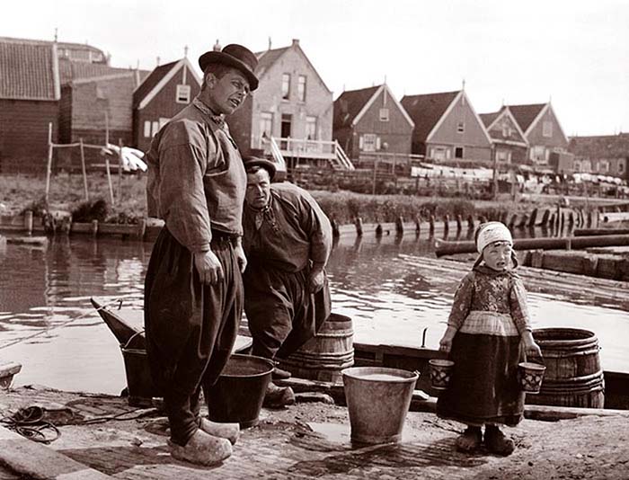 Два рыбака в мешковатых штанах, деревянных башмаках стоят на причале вместе с маленькой девочкой, держащей маленькие ведерца. Маркен, Нидерланды.