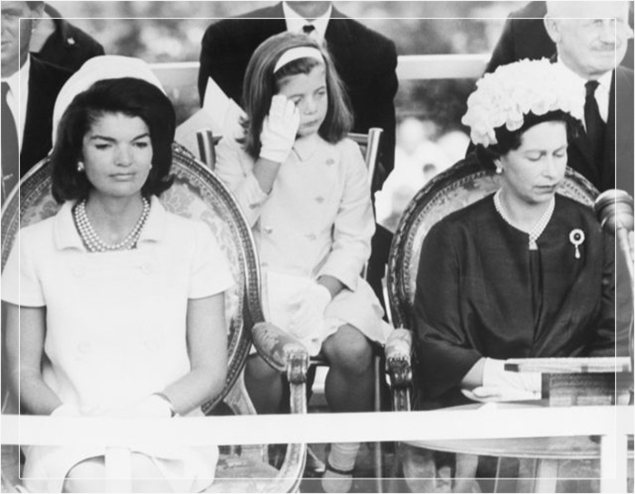 Кэролайн Кеннеди, дочь Джона Ф. Кеннеди и Джеки Кеннеди, вытирает слезу на церемонии, посвящённой мемориалу ее отцу. Перед ней сидят Джеки Кеннеди и королева Елизавета II. 