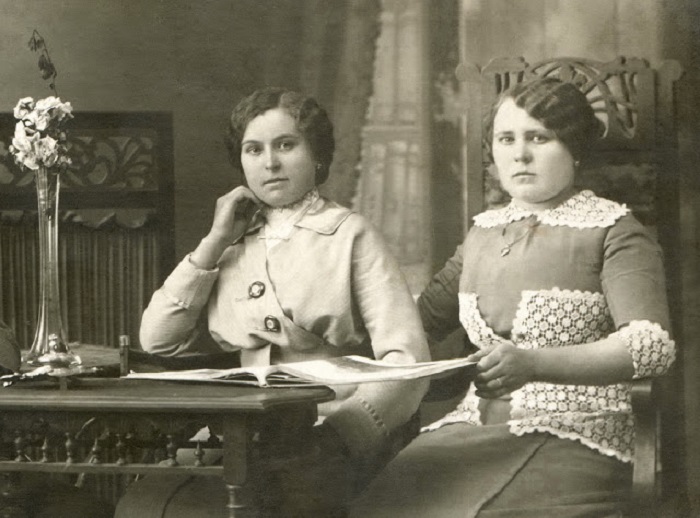 Две сестры читают книгу, сидя за столом. 