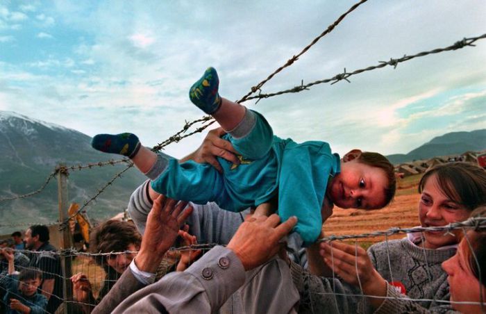 Двухлетнего малыша передают через забор из колючей проволоки в руки бабушки и дедушки на албанской стороне.