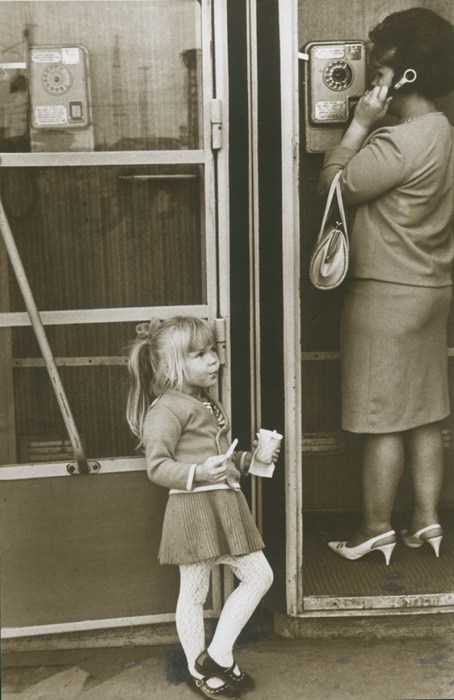 Девочка есть мороженное, пока ее мама разговаривает по таксофону. 