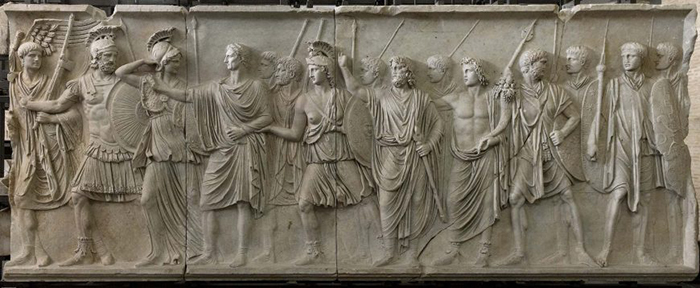 На барельефе когда-то был изображен император Домициан, потом его место занял император Нерва. Изображение преемника (четвертый слева) отличается от остальных фигур неправильным соотношением головы и тела